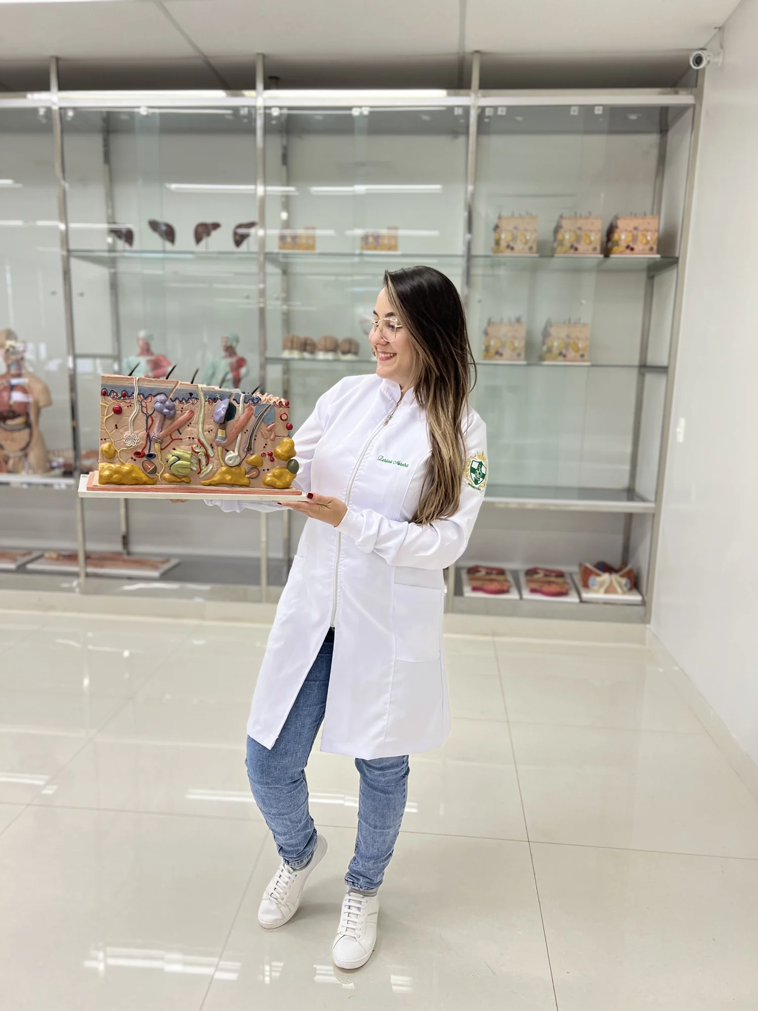 Larissa Moreira, de jaleco branco com o nome bordado e o brasão da Biomedicina na manga, sorri para um modelo anatômico da pele em corte que segura num laboratório de anatomia